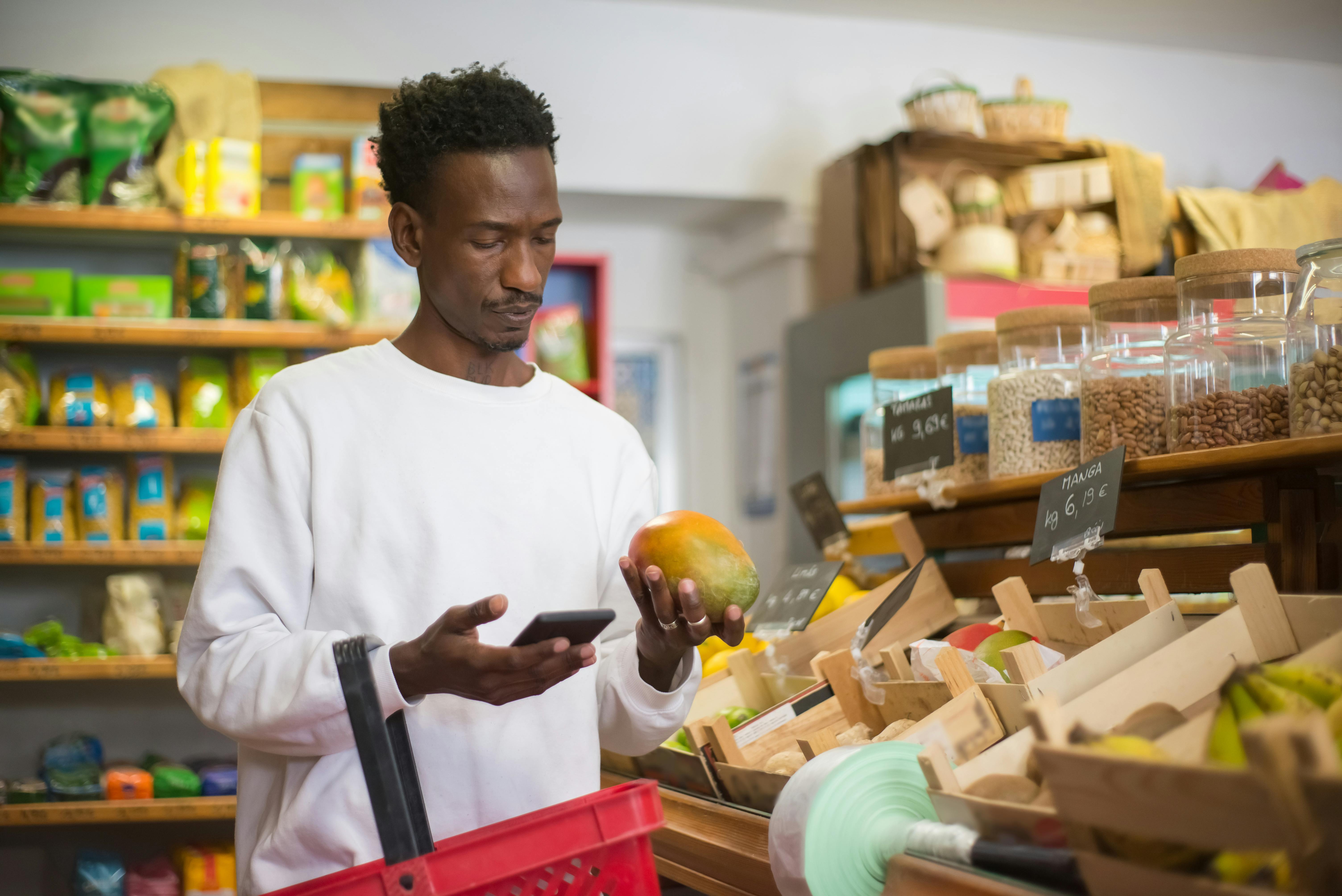 Homem negro segurando uma fruta e um celular comparando preços